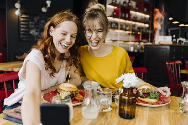 Two happy female friends having burger and taking a selfie in a restaurant